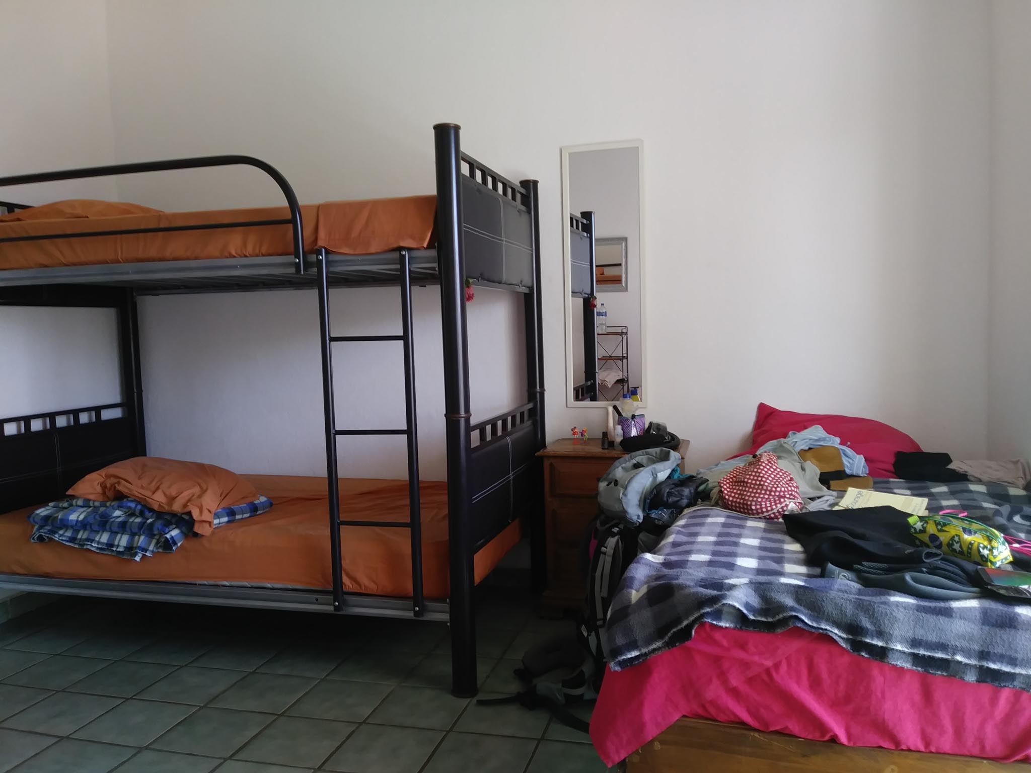 A single bed with pink sheets and someone's belongs strewn all over it is next to a set of black bunk beds with smooth orange sheets.