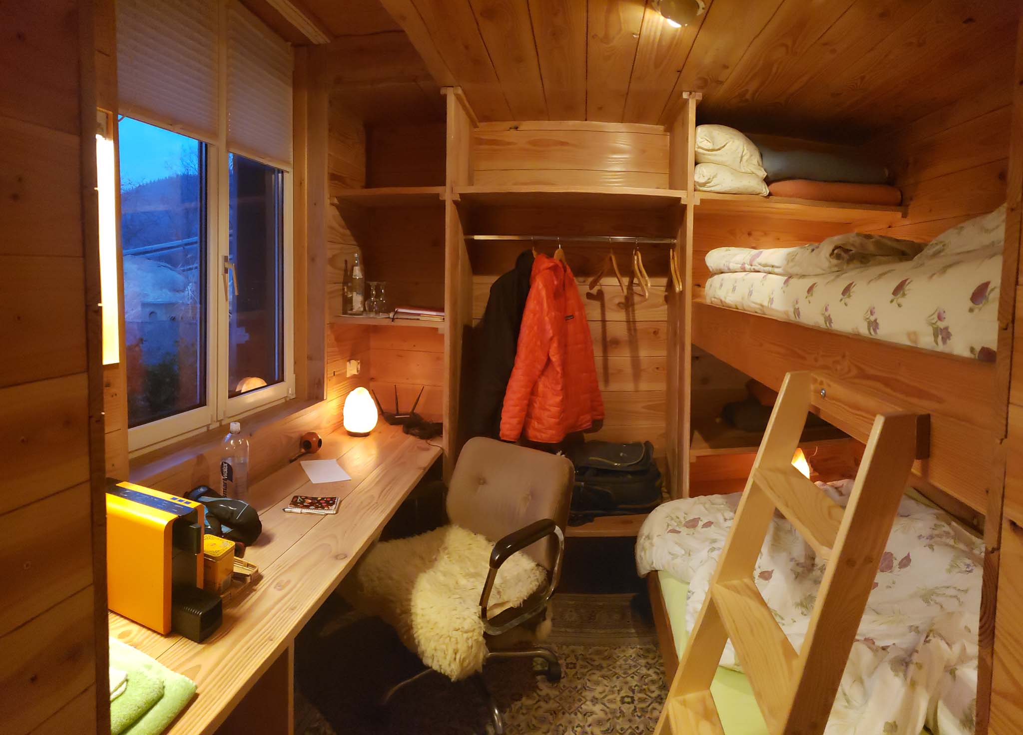 A small, wood lined room shows a bunk bed across from a desk in front of a window.