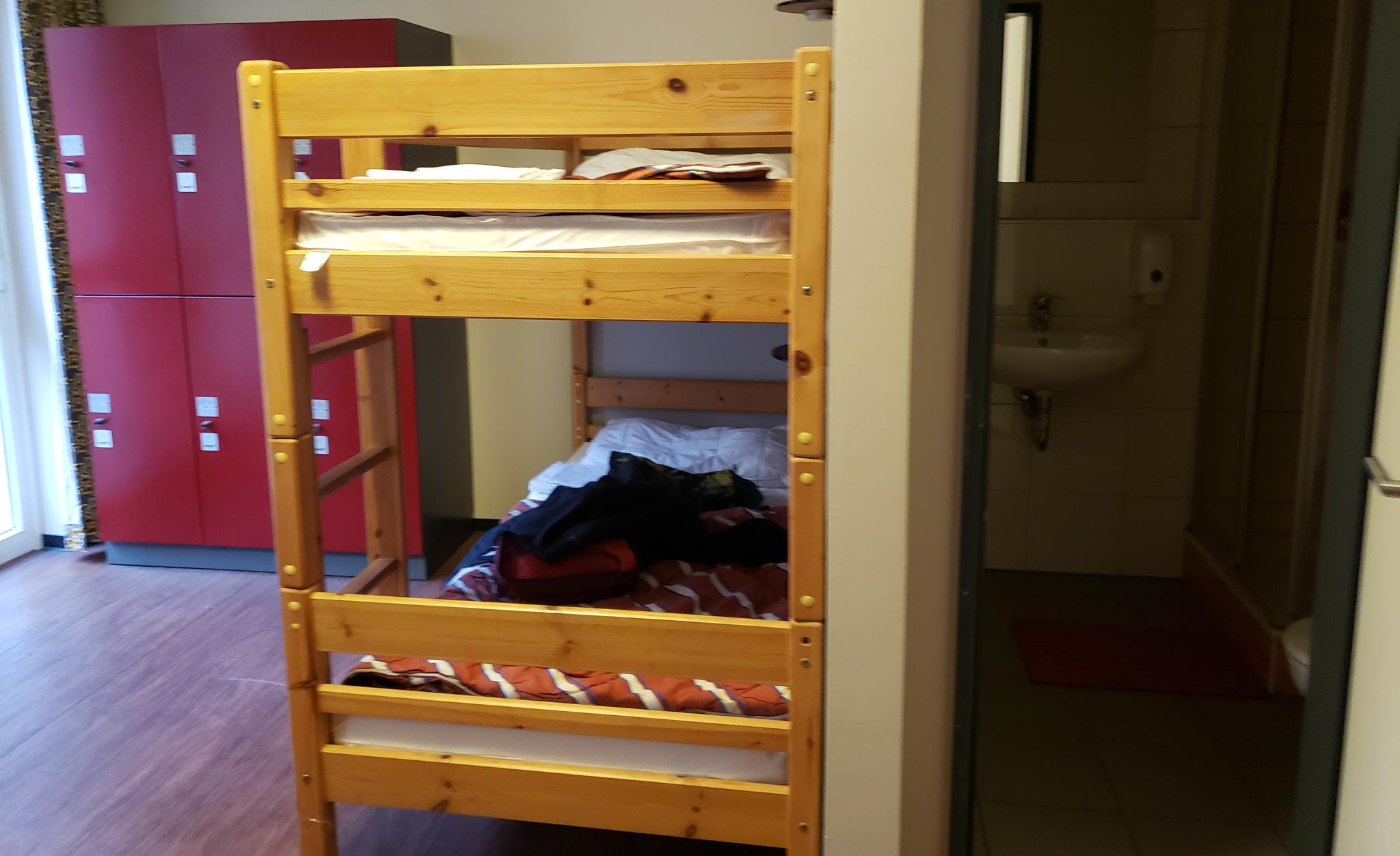 A view of the hostel room shows an open bathroom door, the end of a set of wooden bunk beds with striped orange blankets, and 6 stacked red lockers.