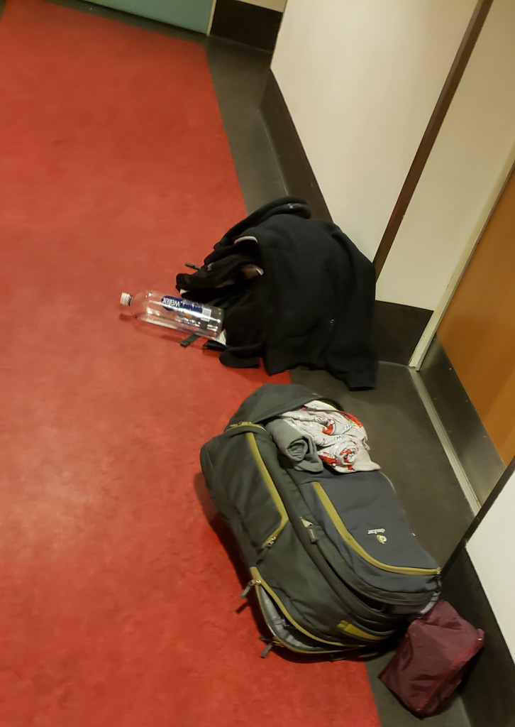 Erin's backpack, laundry, and jacket are piled by a brown door in a hallway with red floors and white walls.