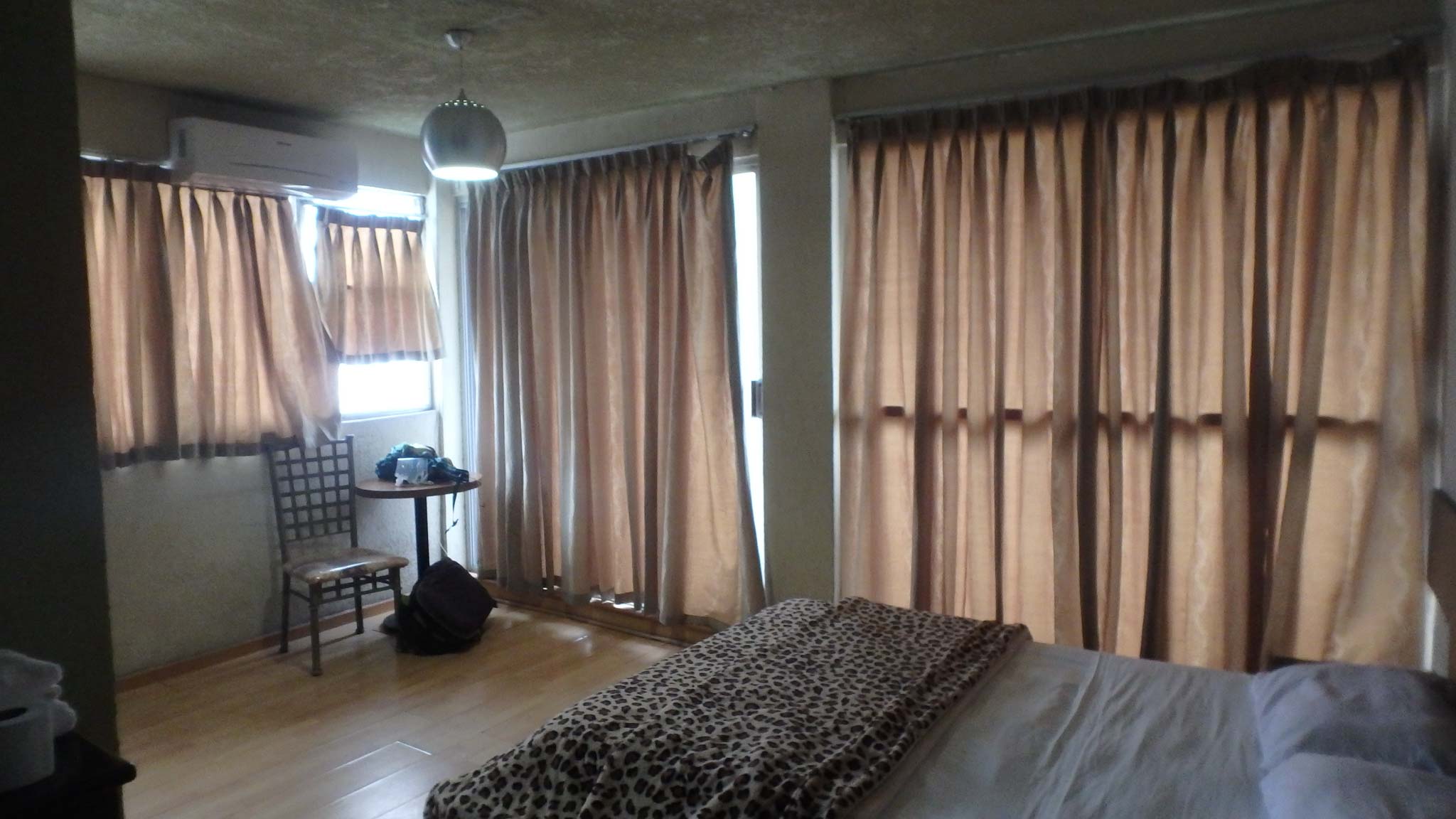 A hotel room with floor to ceiling windows covered with tan curtains and a queen sized bed with white sheets and a leopard spot blanket. Erin's belongings are piled on a small table in the corner.