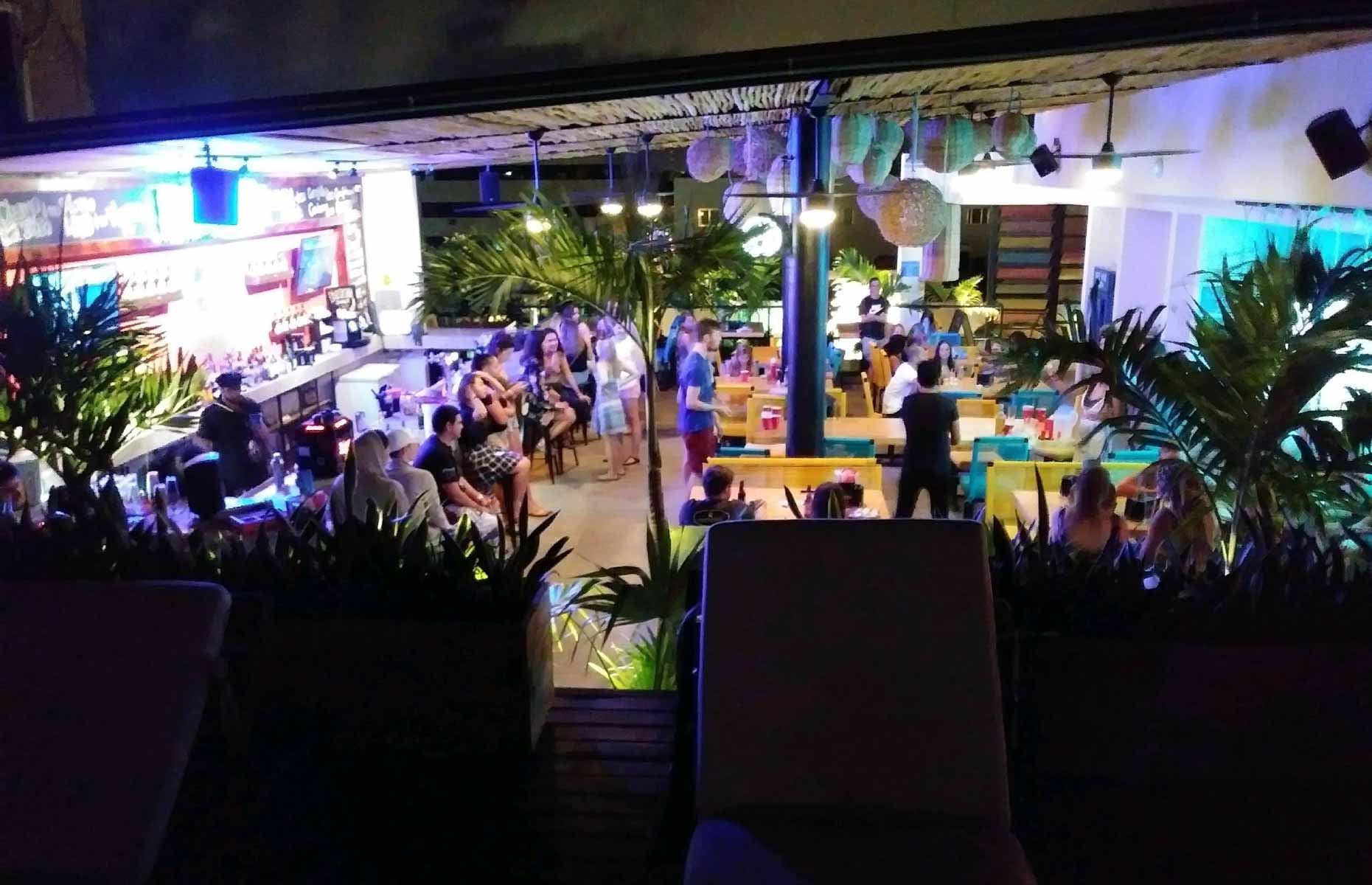 Overlooks a room of green potted trees, tables with bright blue and yellow chairs, and groups of young people watching others play beer pong.
