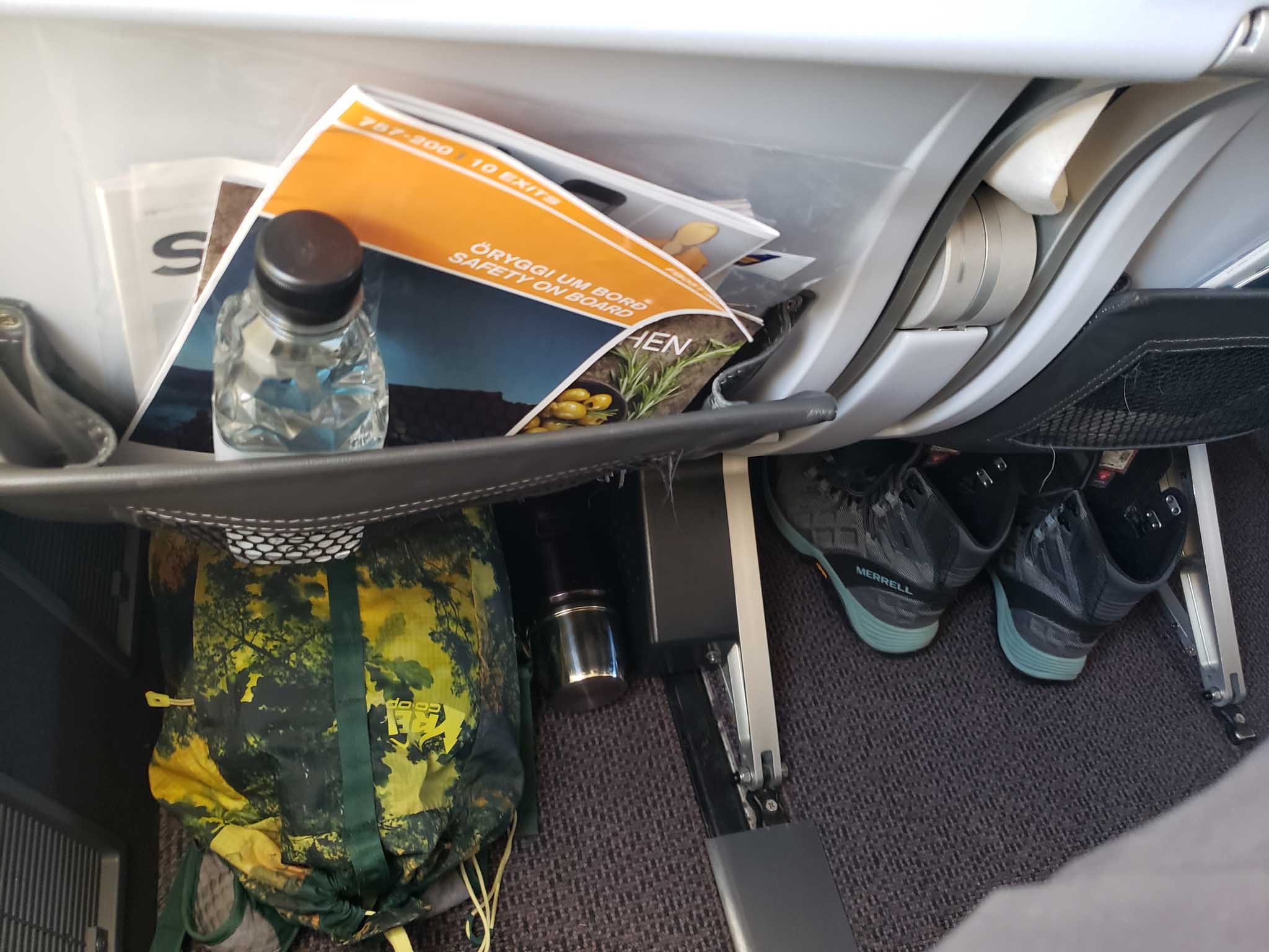 A green and yellow backpack, a black thermos, and a pair of grey and blue boots are pushed underneath two airplane seats.