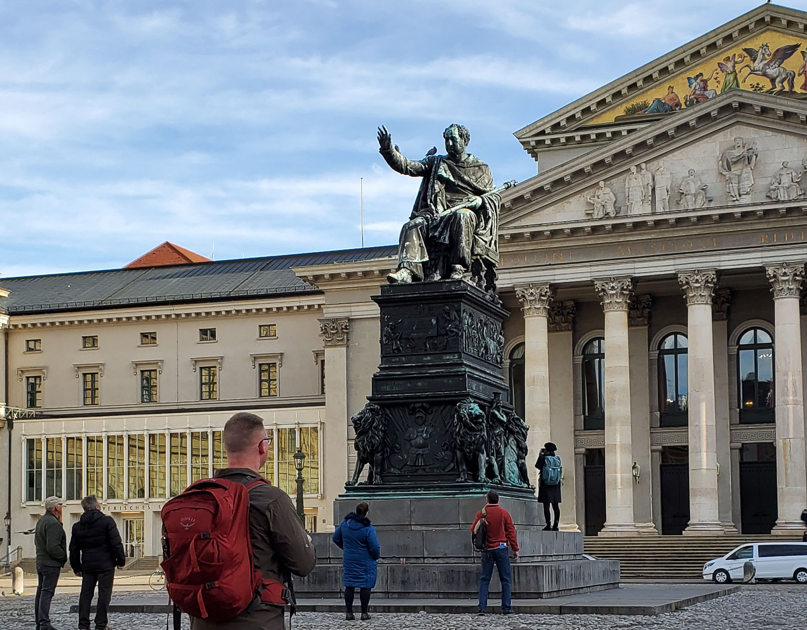 Chris, wearing his red Osprey backpack, looks at a statue of a man sitting with arm outstretch in front of a fancy, old, off-white building in Munich.