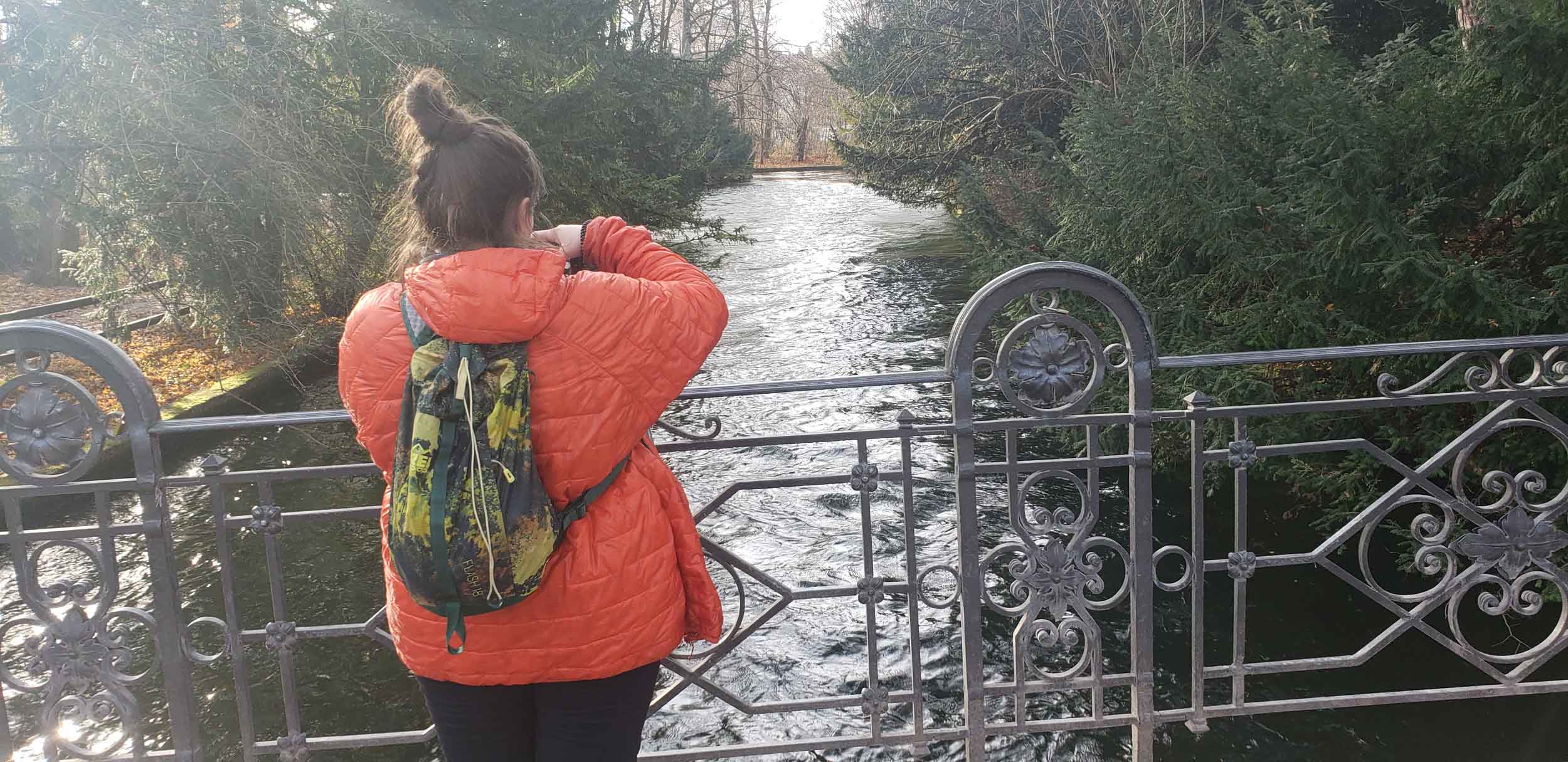 Erin wears a red Patagonia puffy jacket and a small green and yellow REI backpack while shooting photos from a small bridge overlooking a small waterway. Her brown hair is in a very tall, very messy bun.