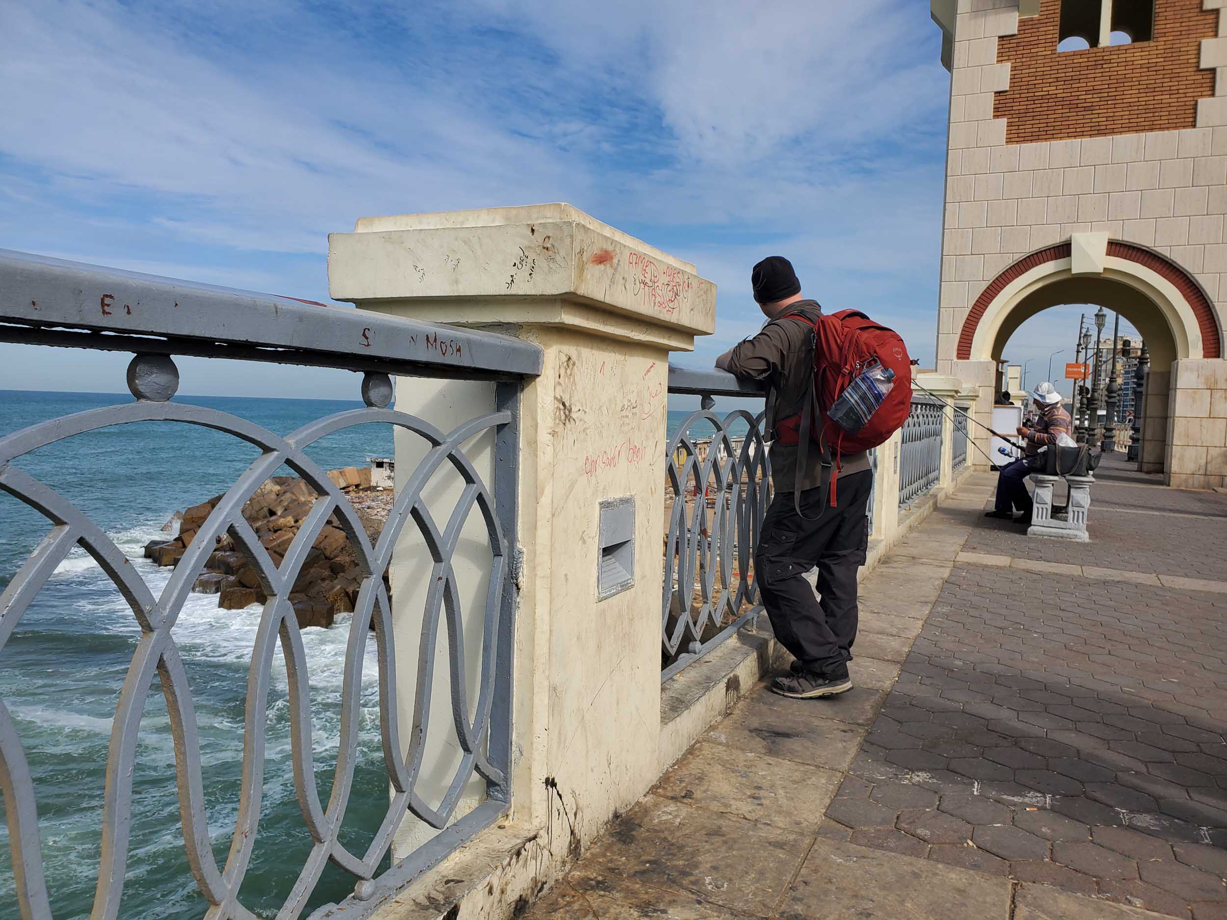 Chris, wearing a red Osprey backpack, stands on a bridge overlooking the Mediterranean Sea and watches an old man in a white sun hat fishing from a bench on the bridge.